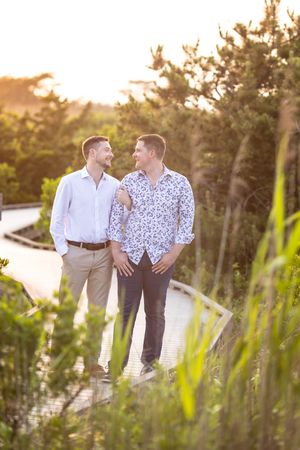 Two men share a loving glance while walking on a sunlit boardwalk path surrounded by lush greenery.