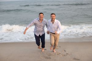 Joyful same-sex couple runs barefoot along ocean shoreline, holding hands and smiling at beach.