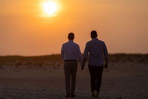 Same-sex couple holds hands walking toward stunning orange sunset on sandy beach at dusk.
