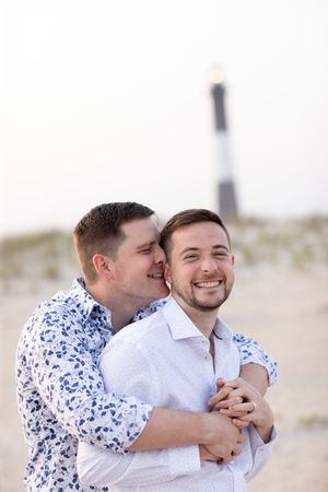 Same-sex couple embraces joyfully on beach with historic lighthouse visible in background at dusk.
