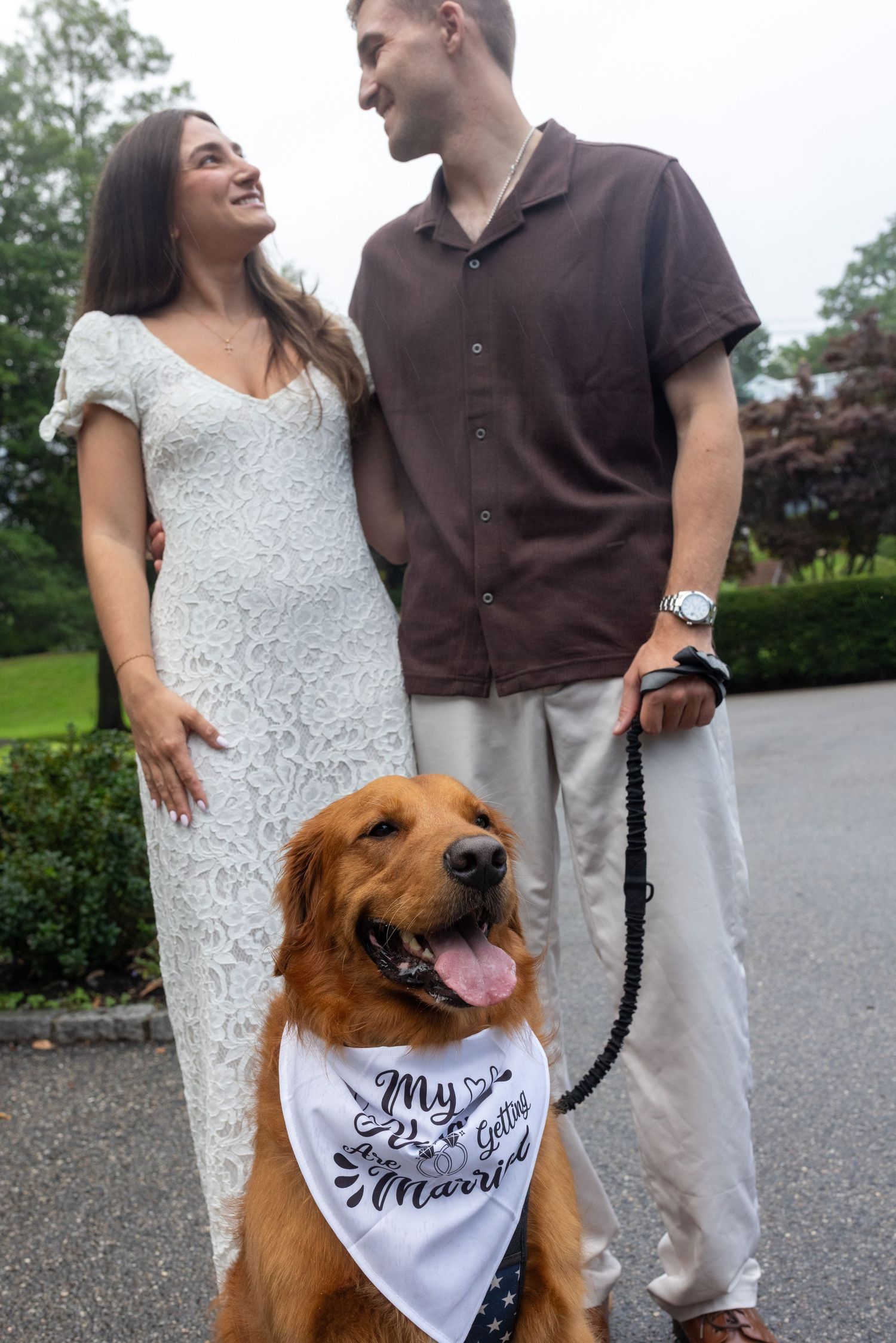 Smiling couple with golden retriever wearing \"My Parents Are Getting Married\" bandana outdoors.