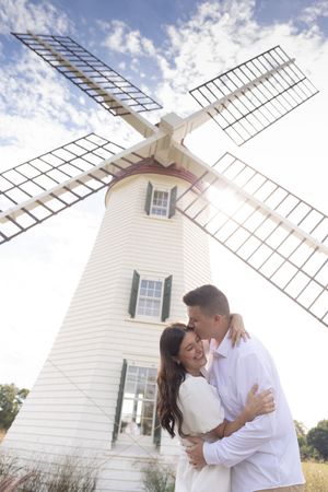 Couple embracing and smiling beneath a white windmill under a bright blue cloudy sky.