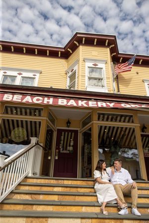 Couple sitting together on steps of charming yellow Beach Bakery building with American flag.