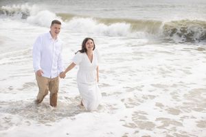 Laughing couple in white outfits holding hands while wading through ocean waves together.