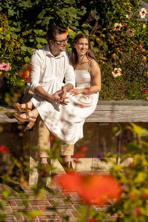 Couple sitting on a stone wall surrounded by colorful flowers in a sunny garden.