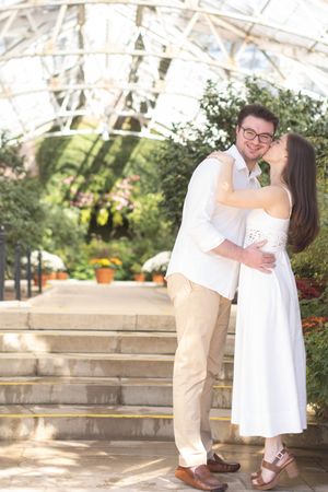 Couple embracing and kissing inside a bright greenhouse with arched glass ceiling.