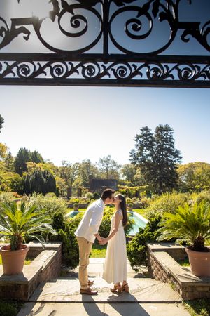 Couple sharing a kiss framed by ornate iron gate overlooking a lush formal garden.