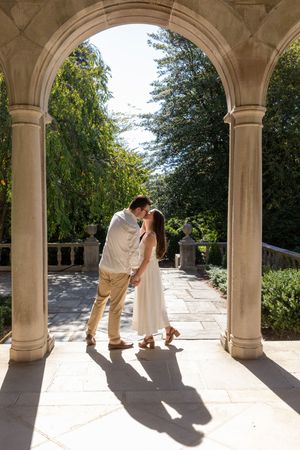 Couple kissing under a stone archway in a sunlit garden colonnade.