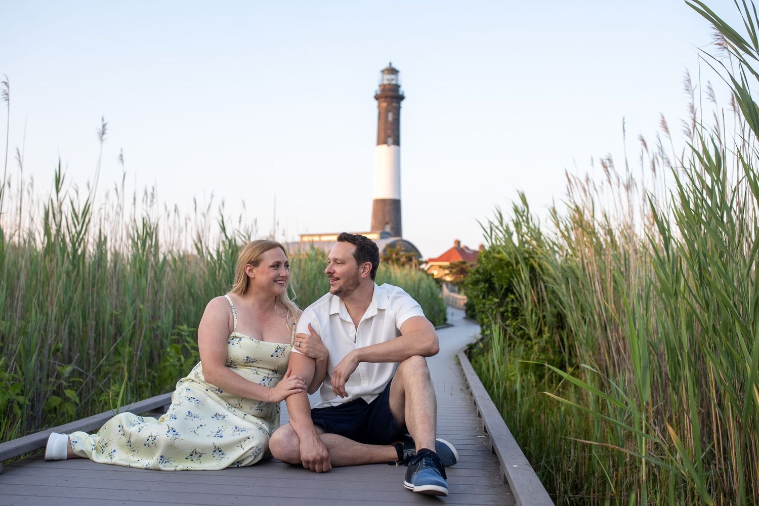 Romantic couple sitting together on boardwalk with historic lighthouse and tall reeds at sunset.