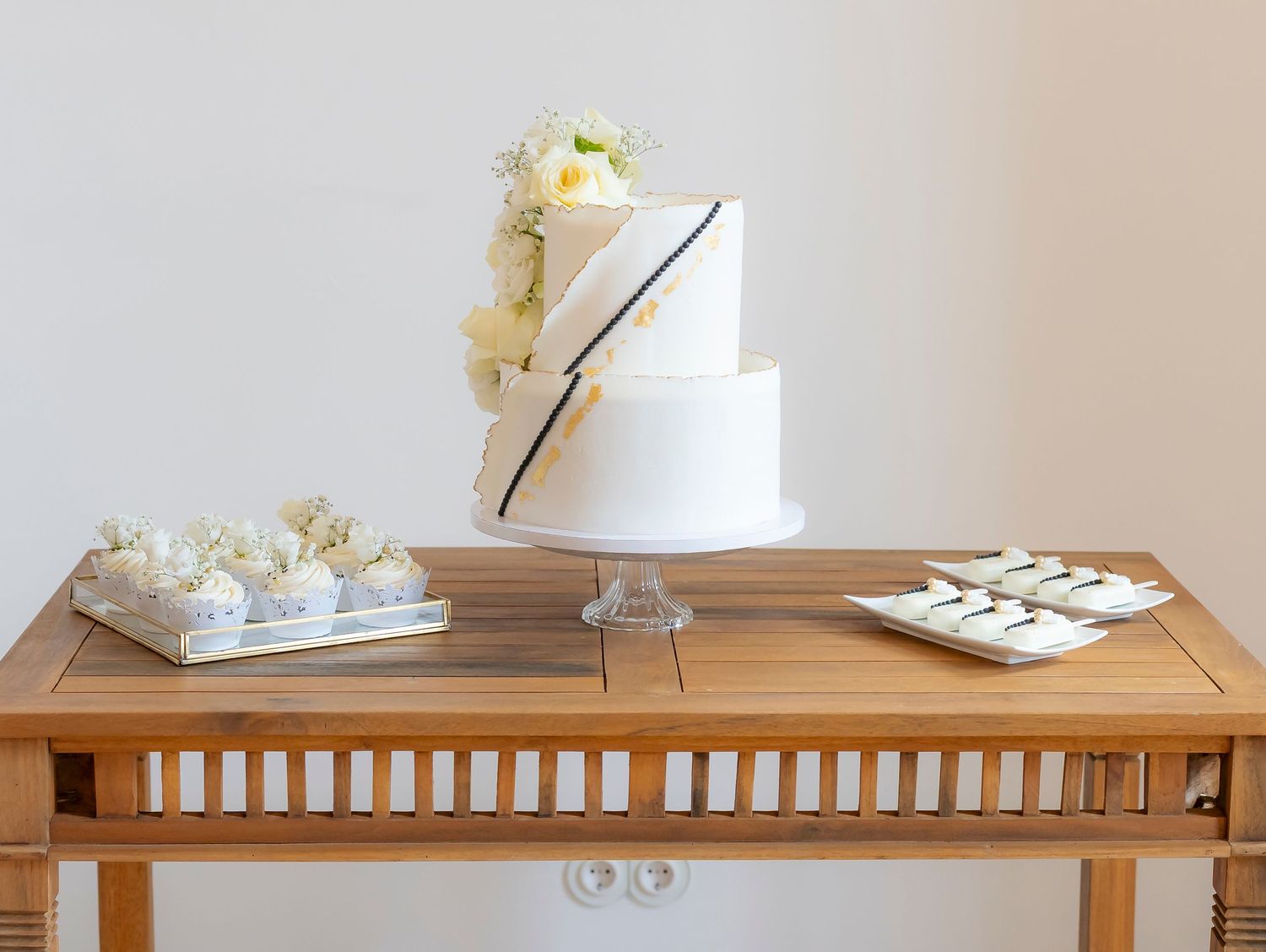 Two-tier white wedding cake with floral accents displayed on wooden table alongside petit four desserts.