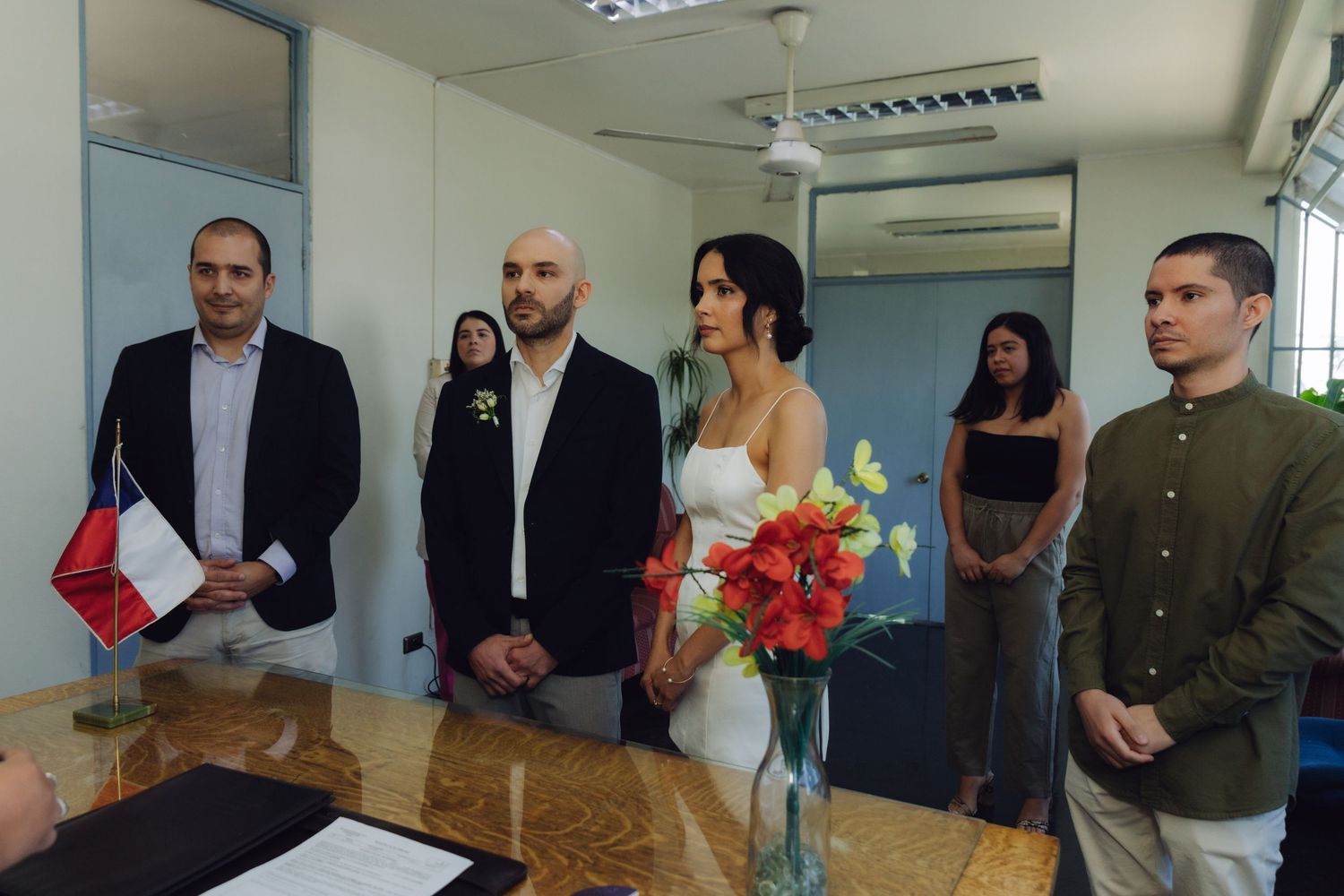 Couple standing at a civil wedding ceremony indoors with guests and a Chilean flag nearby.