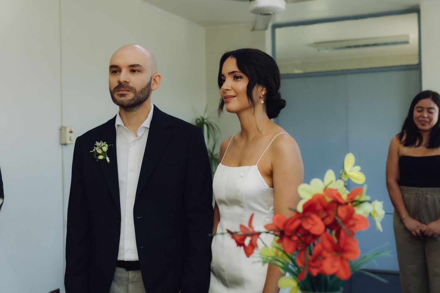 Bride holding red flowers stands beside groom in black blazer during intimate civil wedding ceremony.