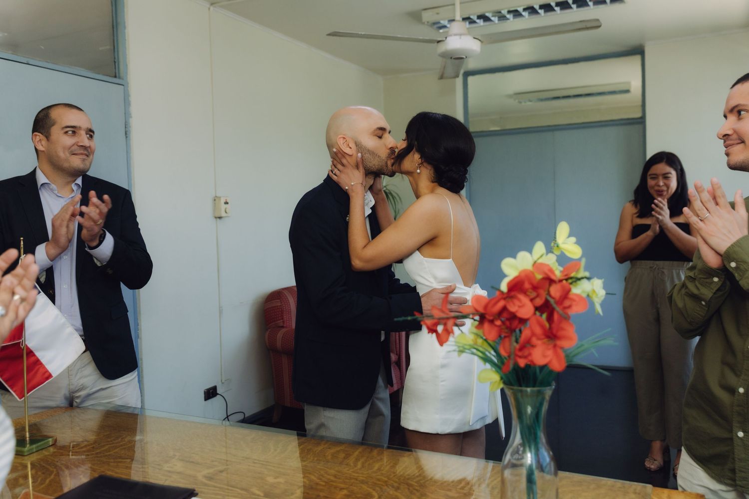 Newlywed couple shares first kiss surrounded by applauding guests at civil wedding ceremony.
