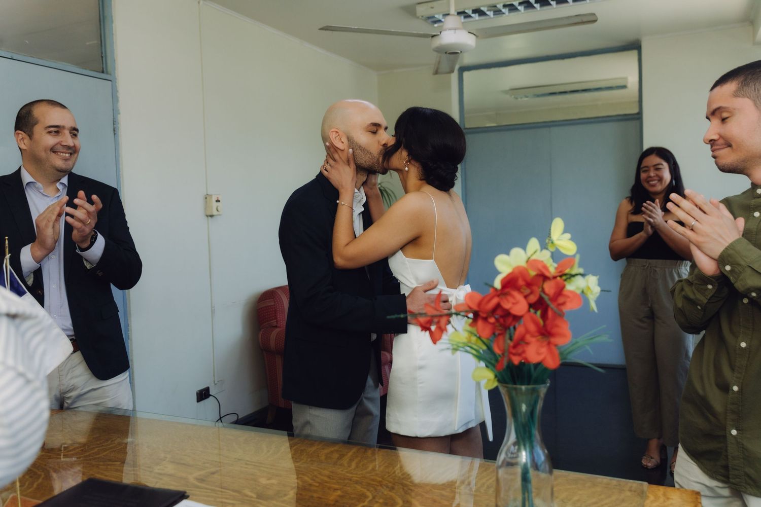 Newlyweds share romantic kiss while wedding guests clap in celebration at civil ceremony.