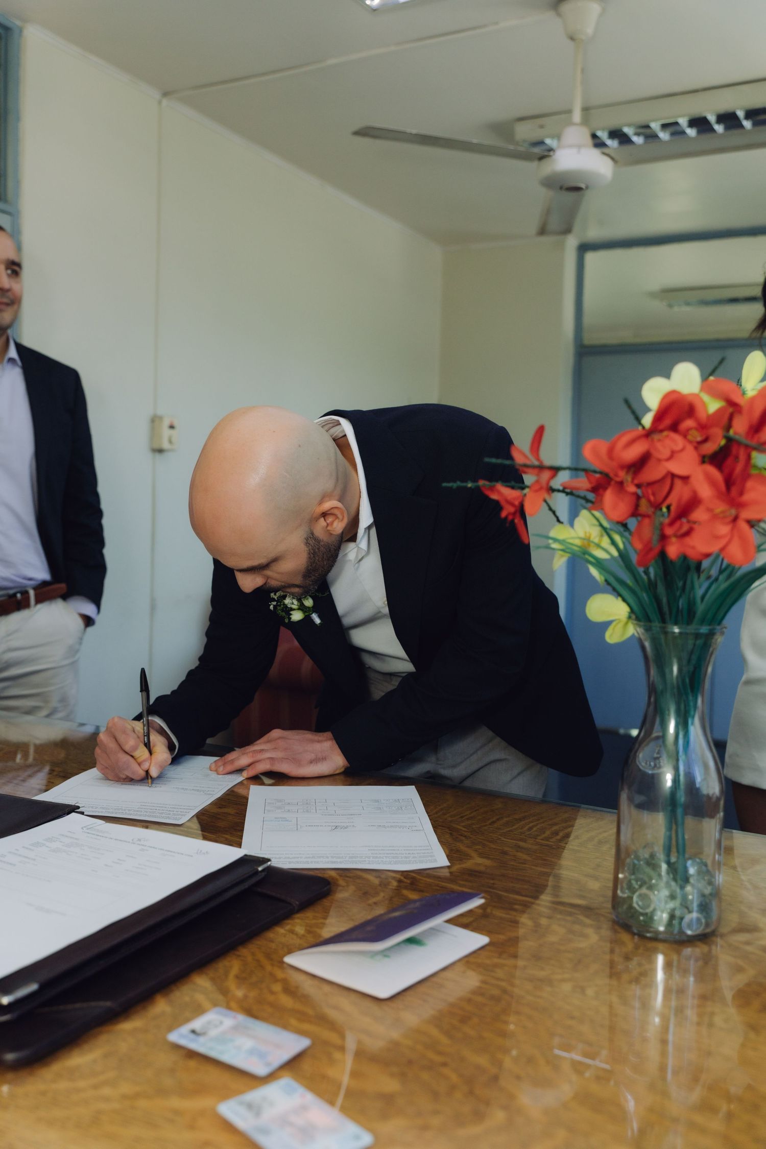 Groom in black blazer signs official wedding documents at wooden table with red flowers nearby.