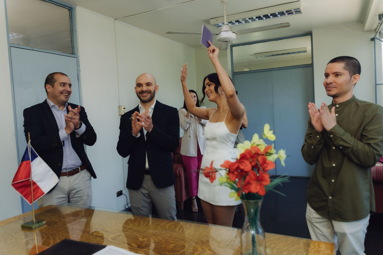 Bride raises marriage certificate triumphantly as groom and guests applaud at civil wedding.