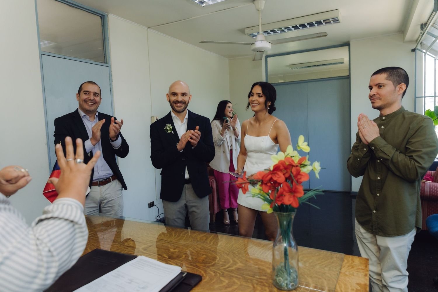 Smiling wedding party applauds newlyweds during intimate civil marriage ceremony indoors.