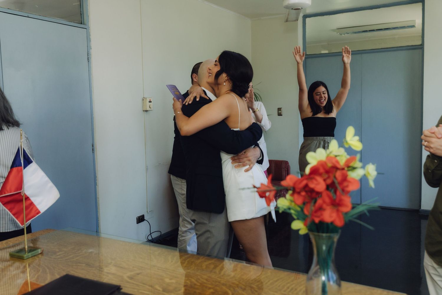 Bride and groom embrace warmly as excited guest celebrates in background at wedding ceremony.