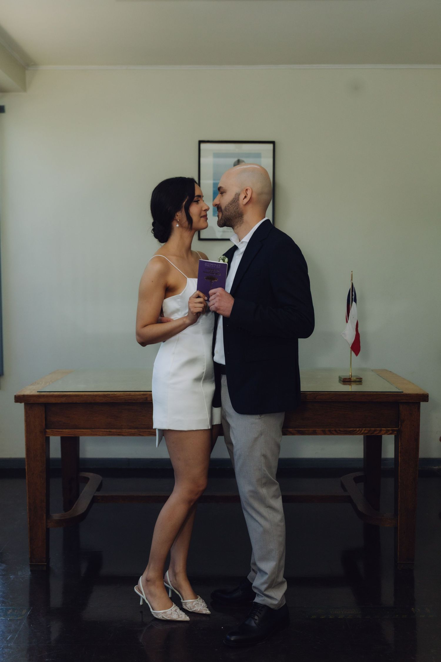 Couple in wedding attire gazing at each other holding a document in a civil ceremony room with Chilean flag.
