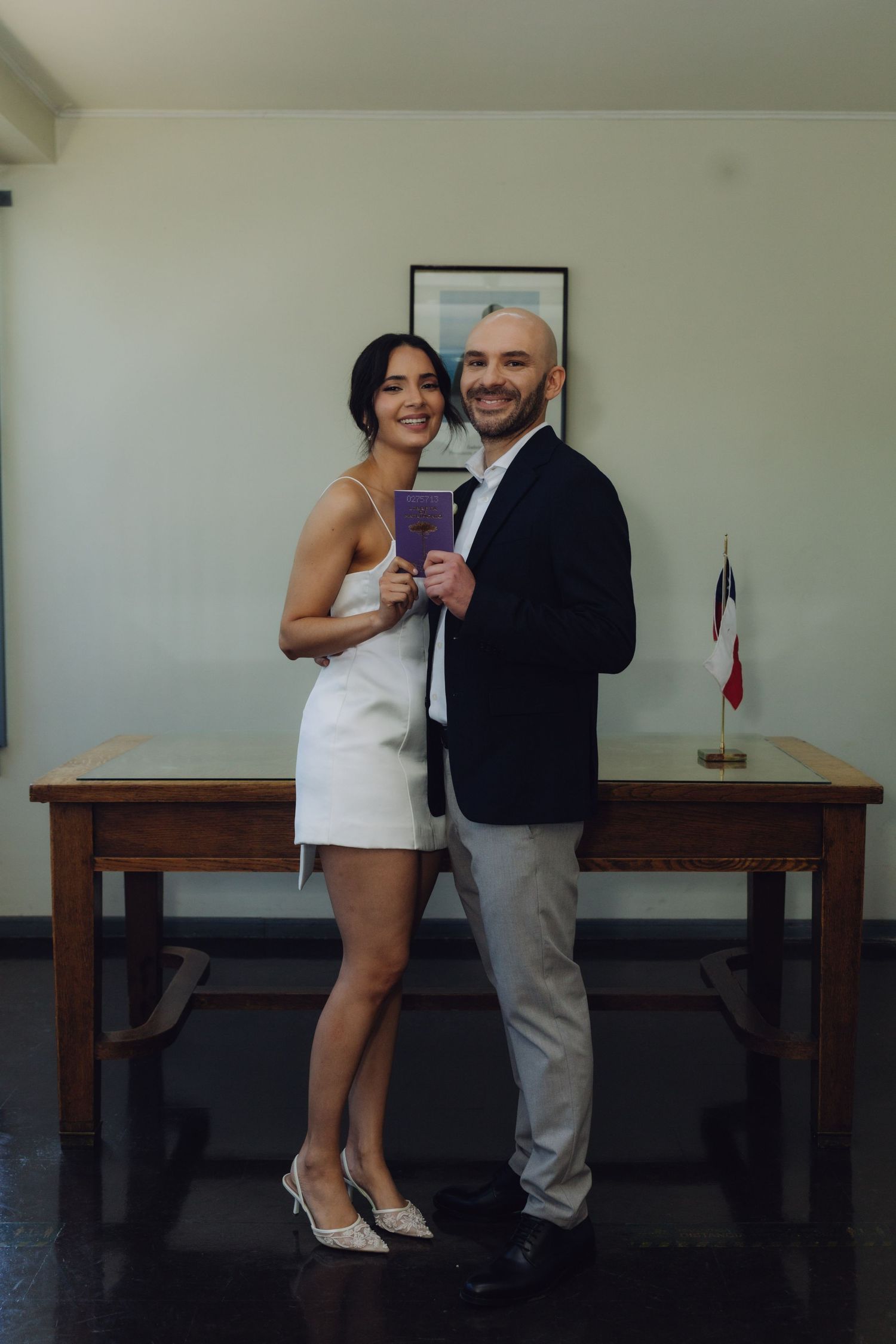 Smiling newlywed couple posing together holding marriage certificate in official ceremony room with flag.