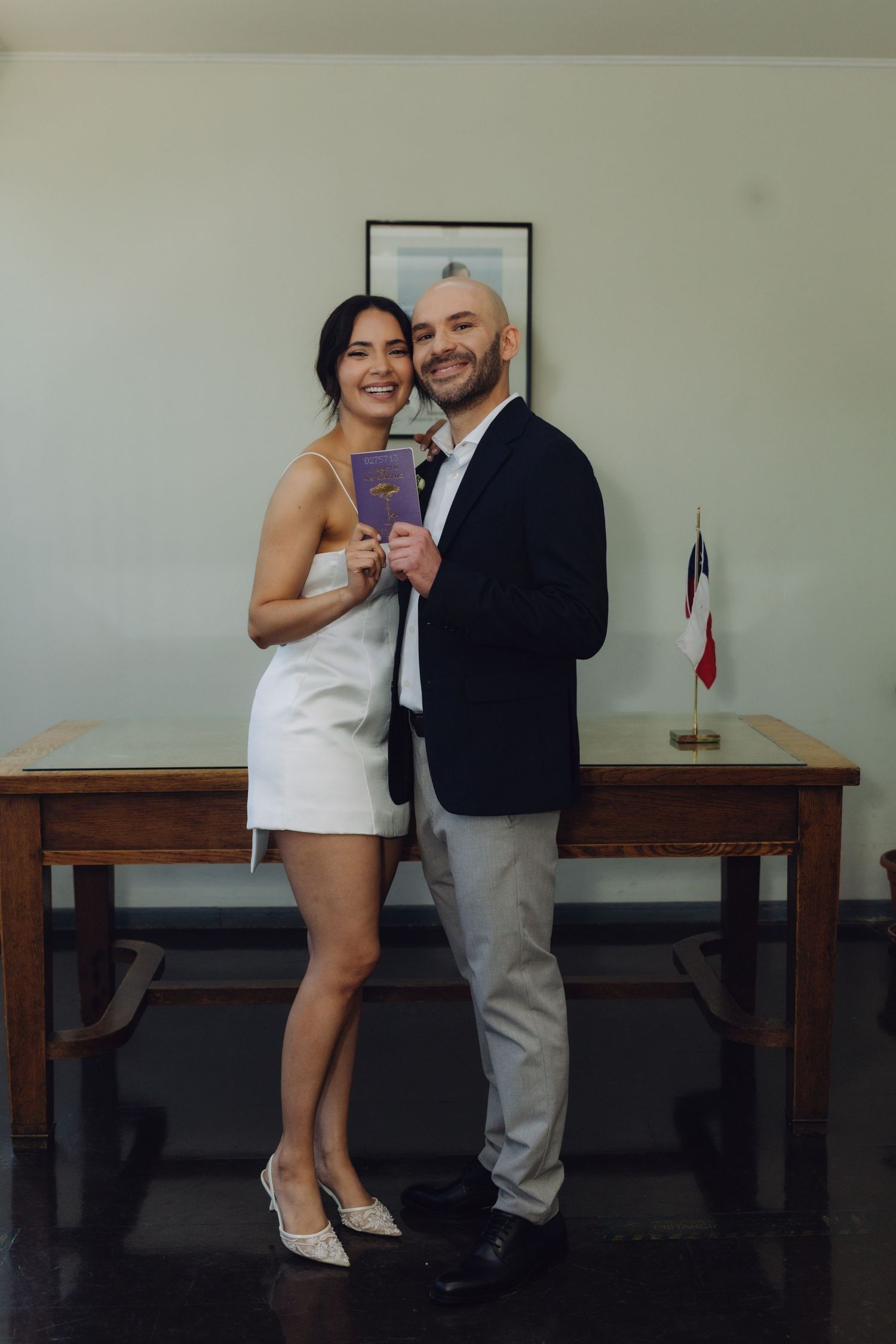 Happy couple in wedding outfits holding marriage document standing before wooden table with Chilean flag.
