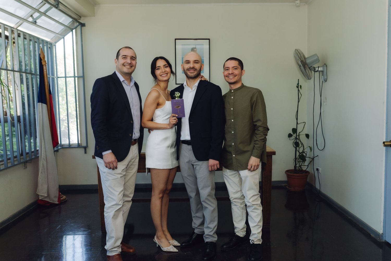 Bride in white mini dress and groom posing with two male guests at civil wedding ceremony.
