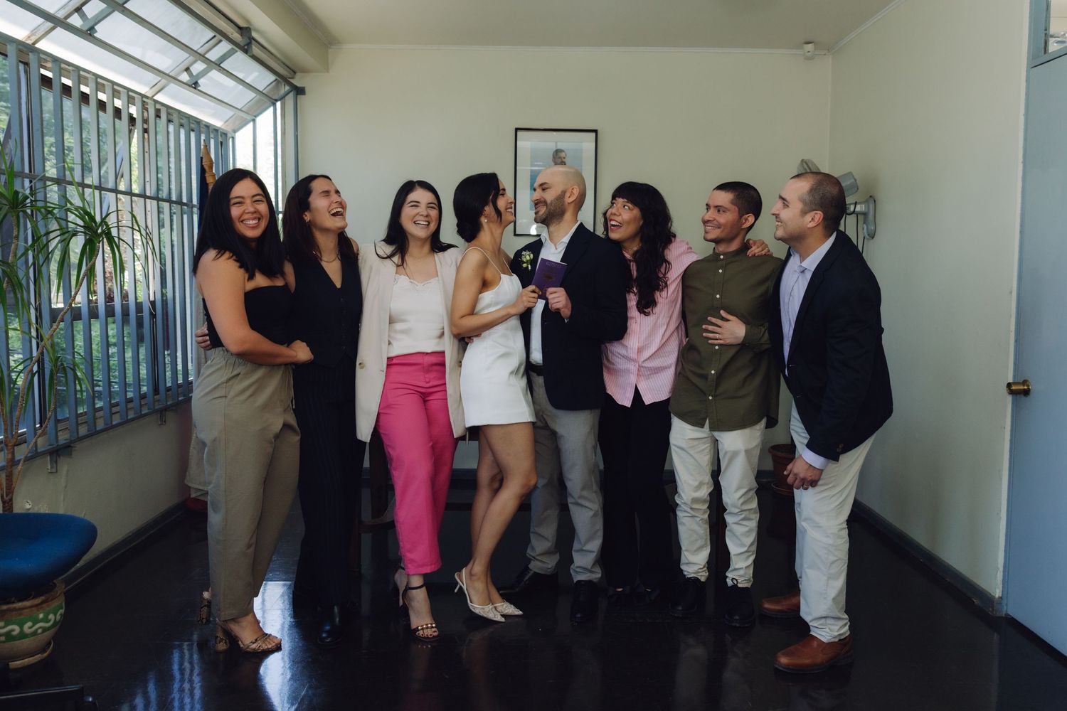 Joyful wedding party group of eight guests celebrating with bride and groom in bright indoor venue.