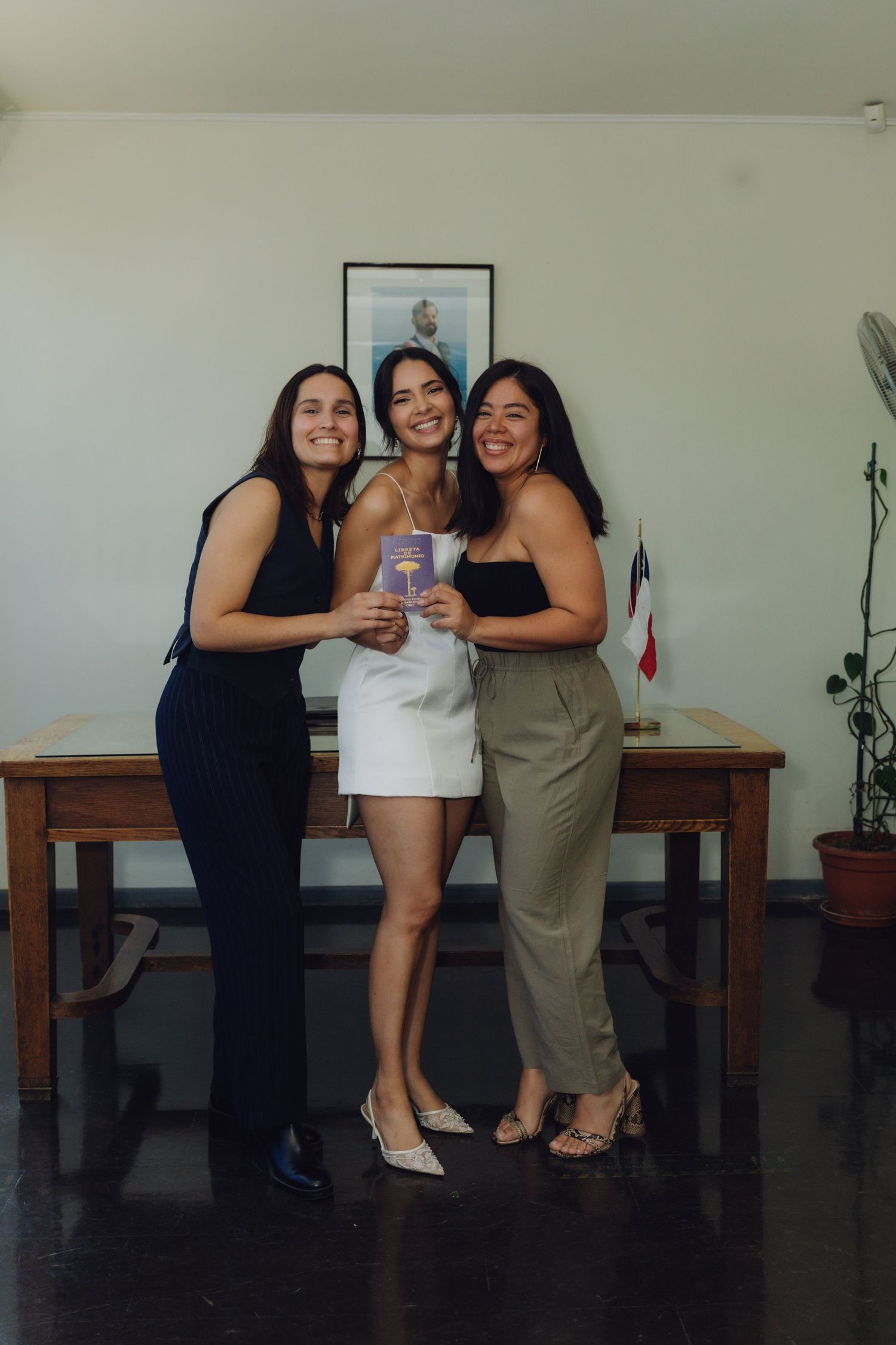Bride in white dress holding marriage certificate smiling with two female friends at civil wedding.