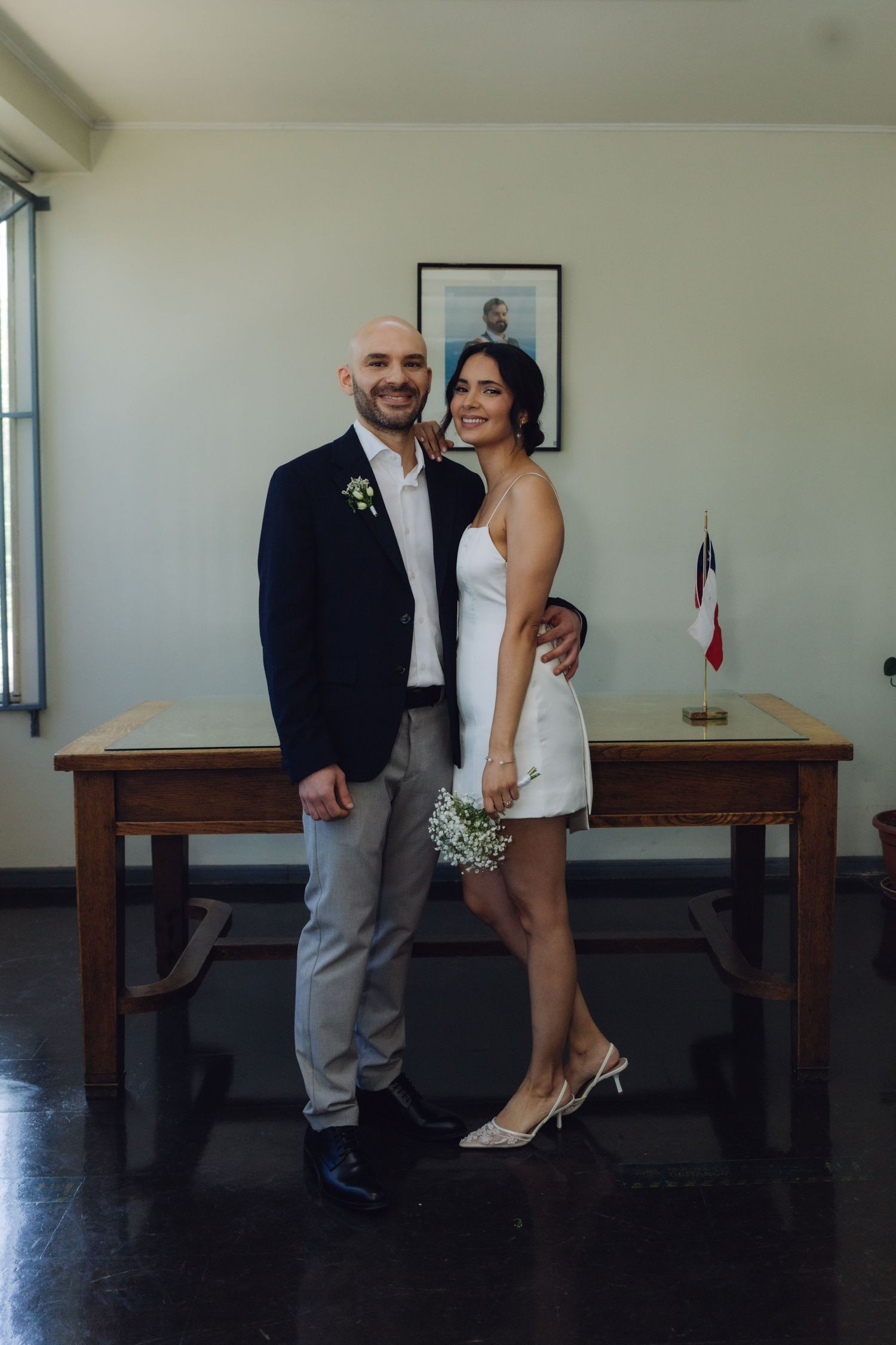 Smiling groom in navy blazer and bride in white mini dress holding flowers at civil ceremony.