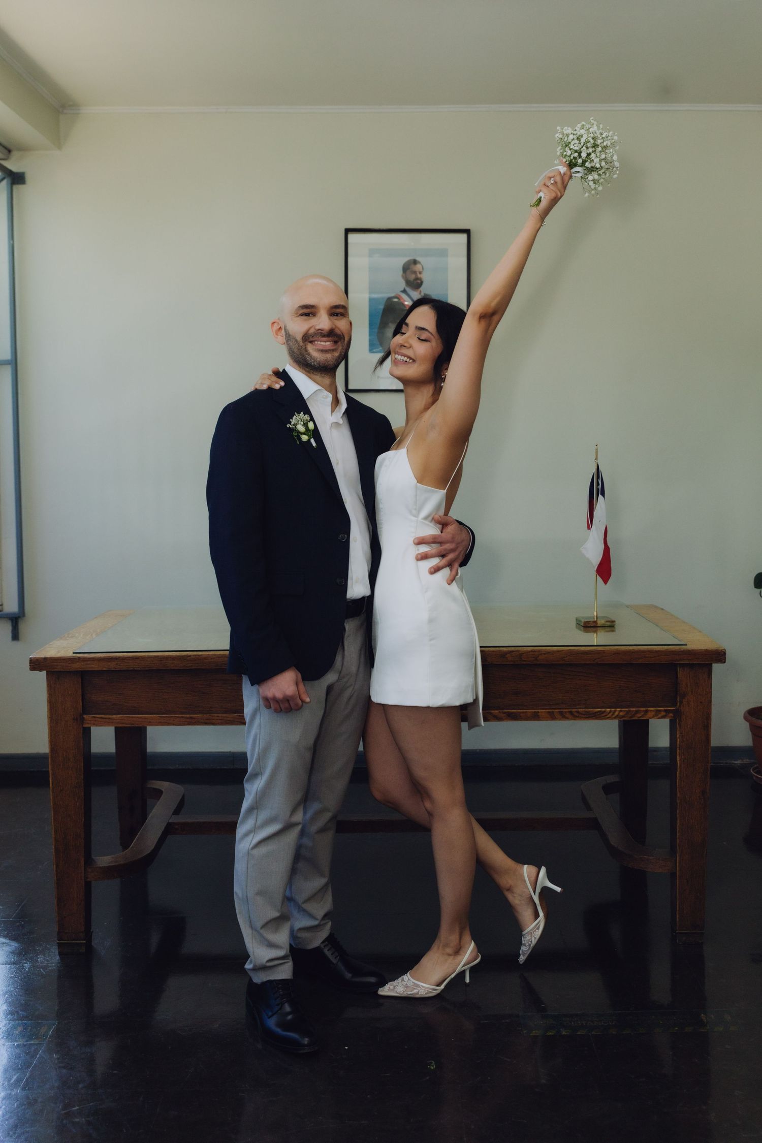 Joyful newlyweds pose after civil wedding ceremony, bride in white mini dress raises bouquet, groom in navy blazer.