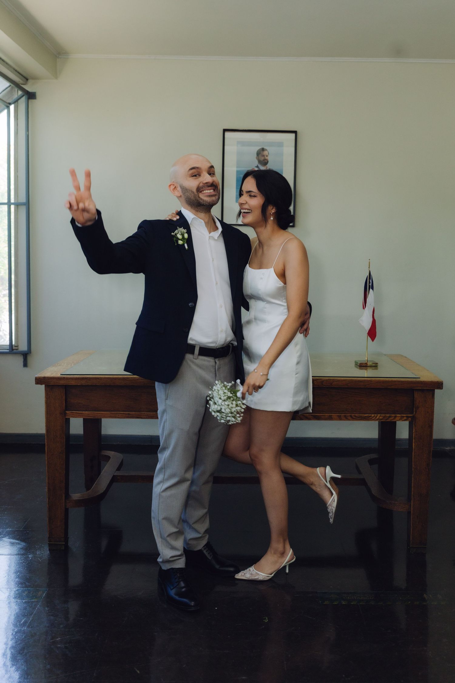 Happy couple celebrates civil wedding, groom flashing peace sign, bride in white dress holding small bouquet.