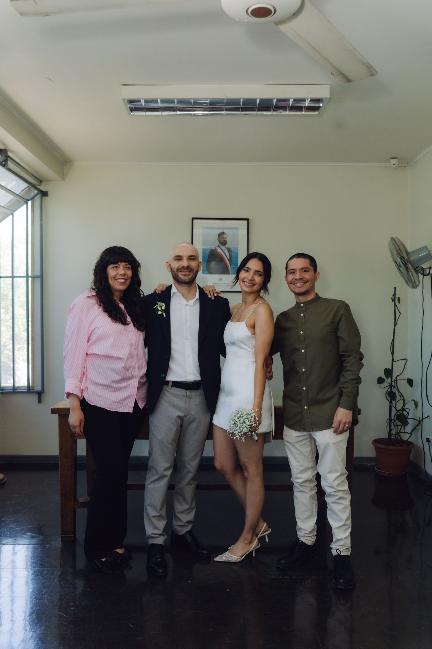Newlyweds and two guests smile together in civil ceremony room with framed portrait on wall behind them.