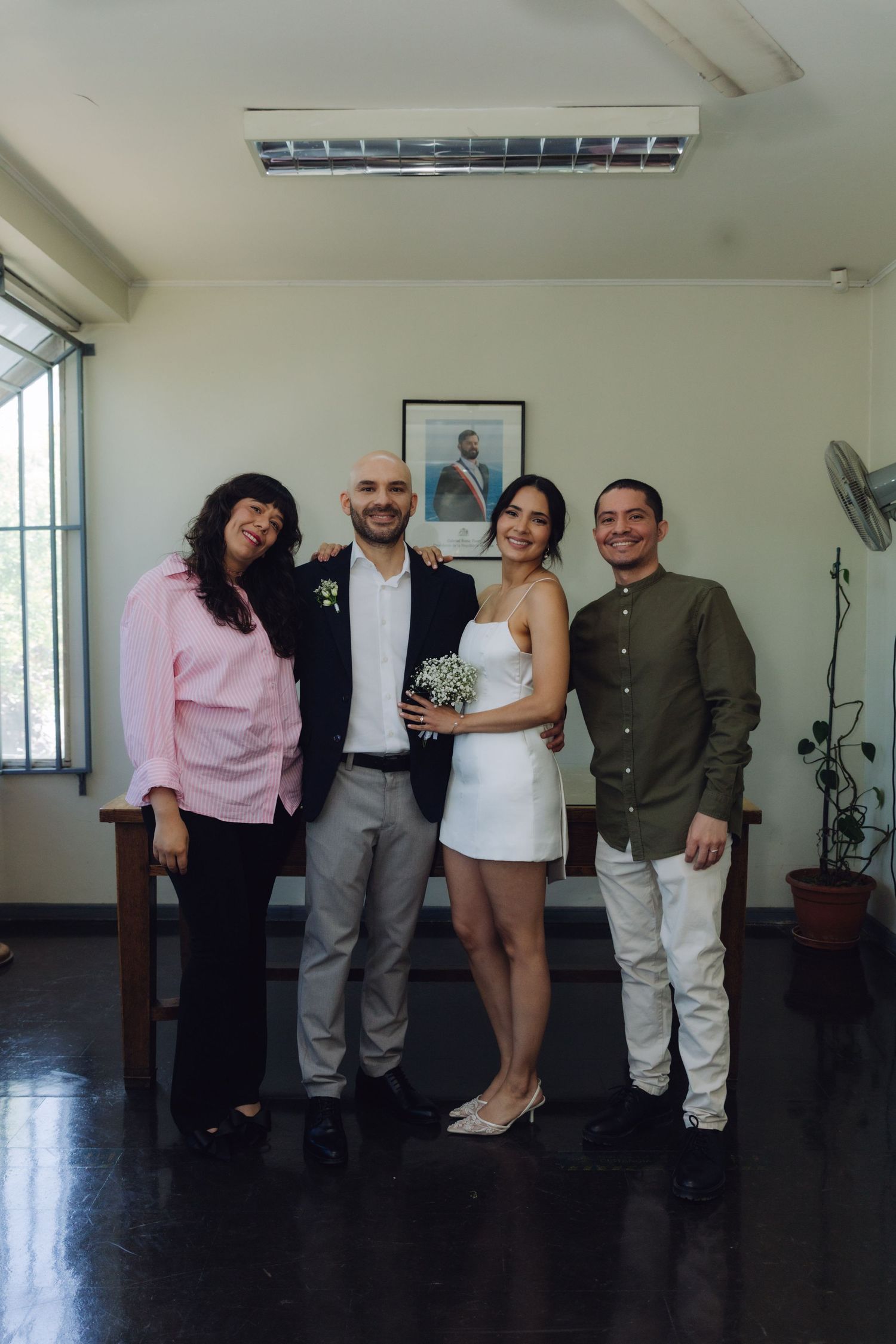 Wedding party of four poses indoors, bride in white mini dress holds bouquet flanked by guests.