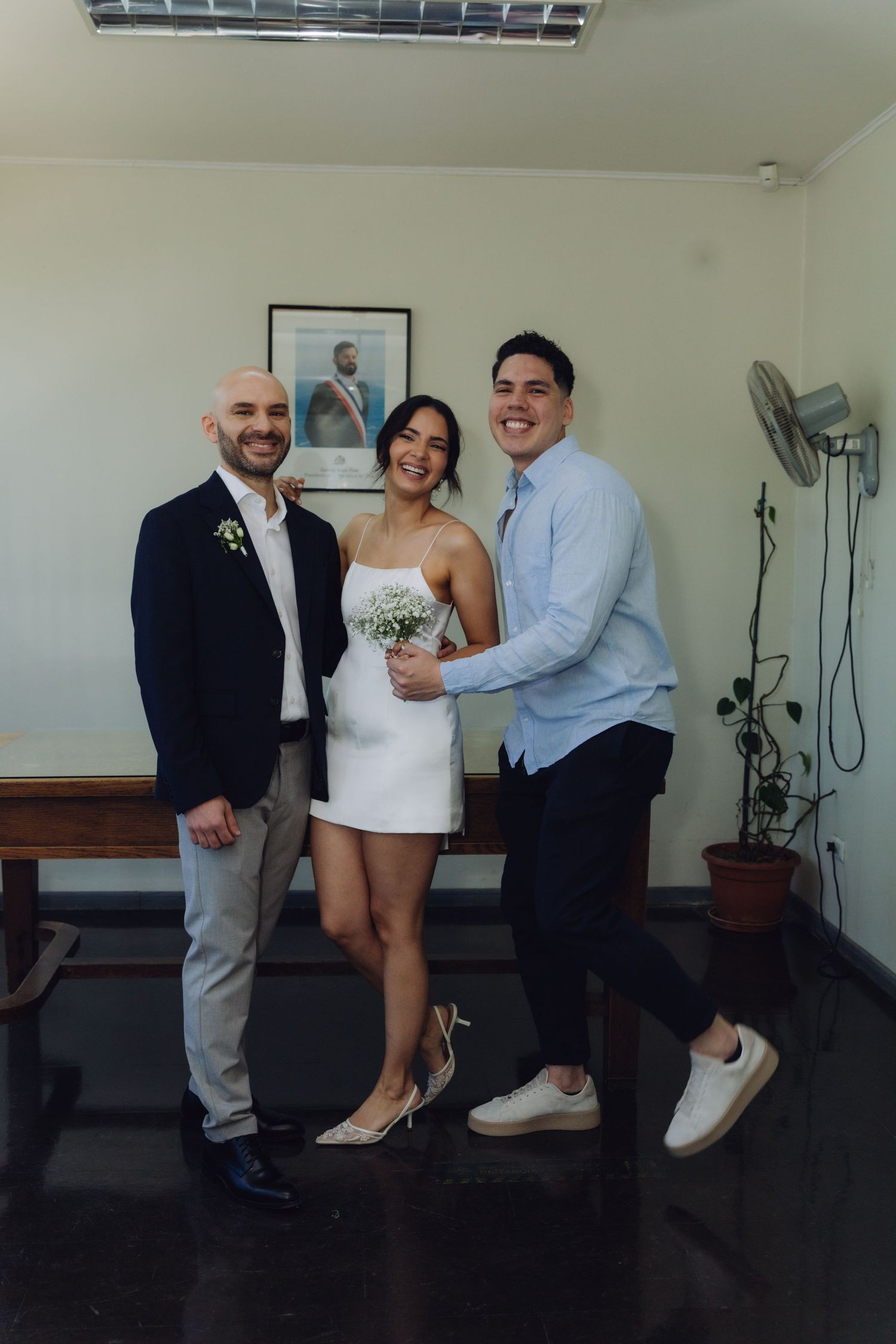 Groom in navy blazer and bride in white dress pose with smiling male guest at civil wedding ceremony.