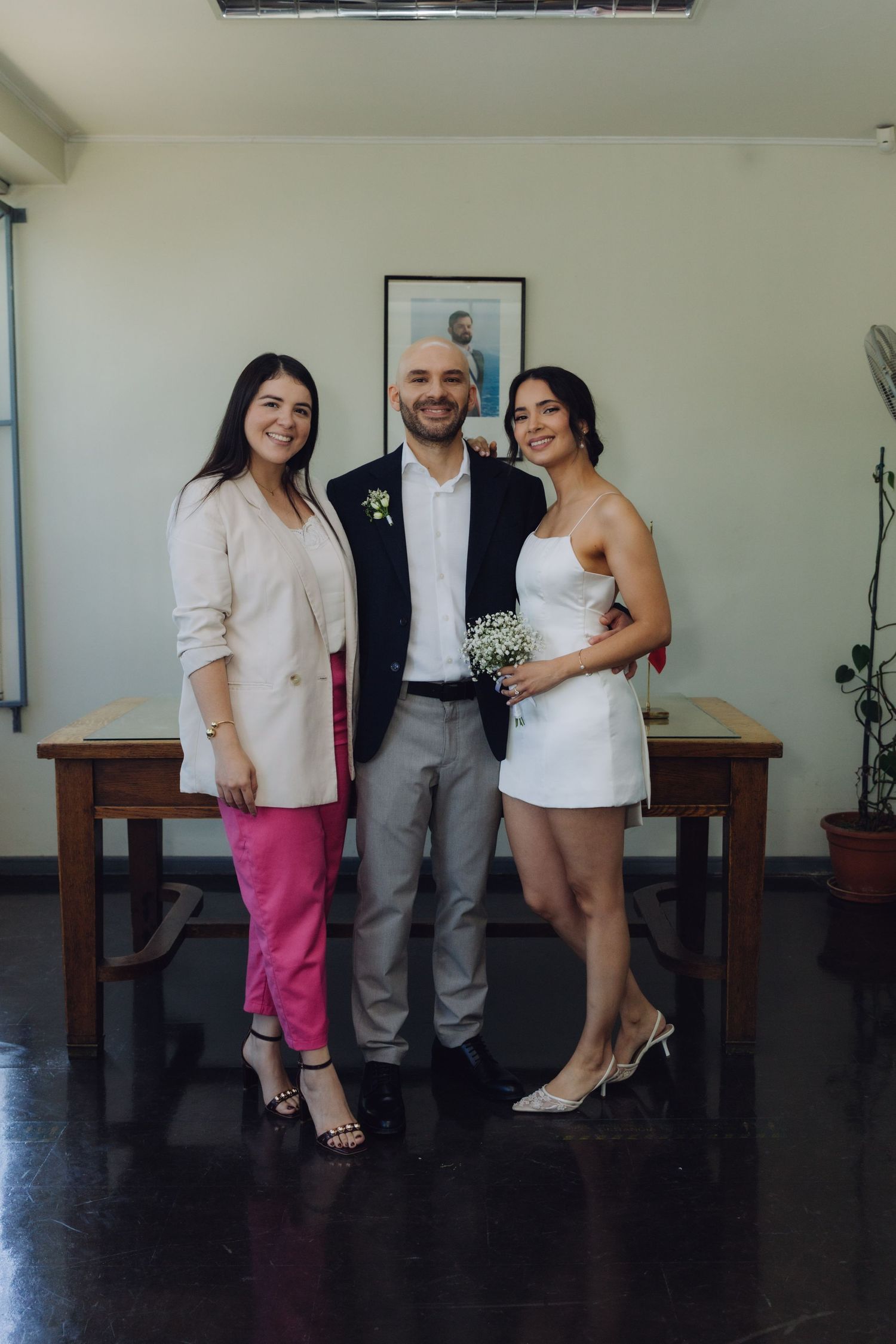 Bride in white mini dress and groom in navy blazer pose with female guest in white blazer and pink pants.