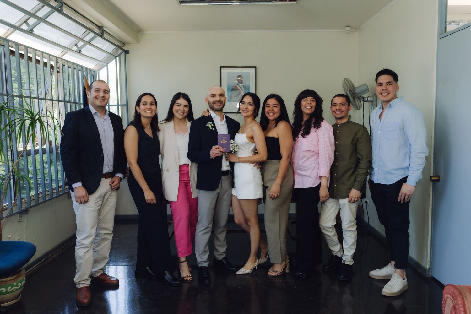 Wedding group photo with nine guests surrounding bride in white dress and groom in navy blazer indoors.