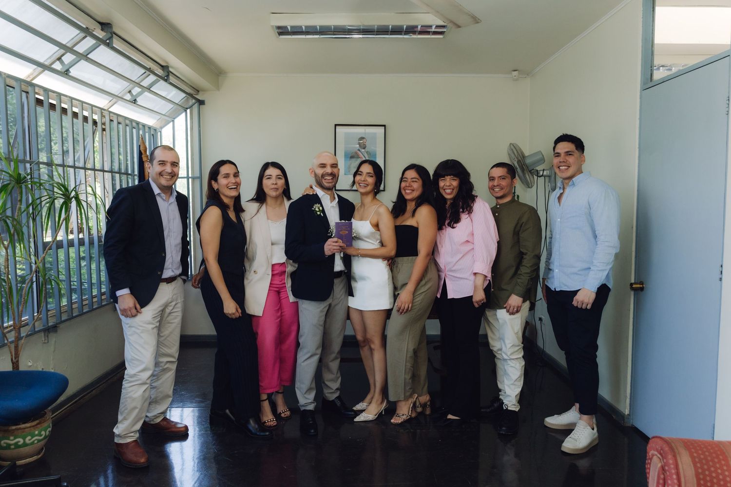 Wedding party of nine people posing together indoors, bride in white dress centered among smiling guests.