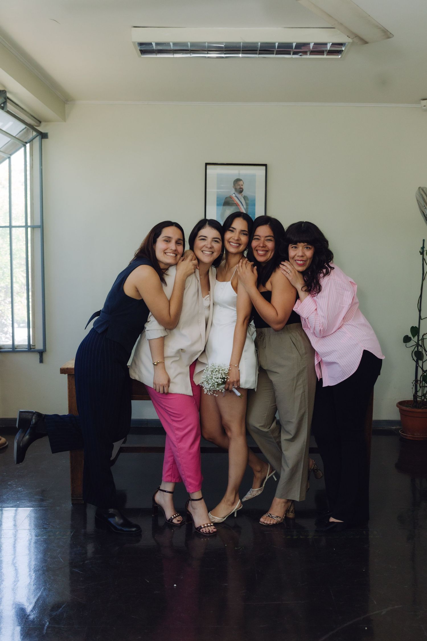 Bride in white dress holds bouquet surrounded by four female friends hugging her at wedding celebration.