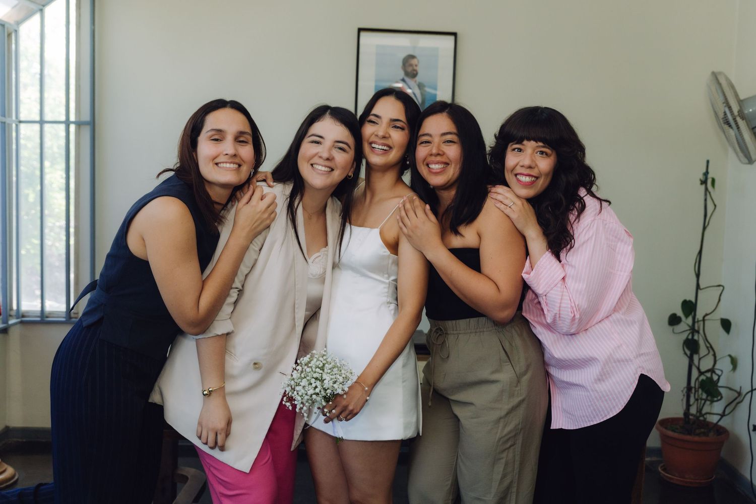 Bride in white dress holds small bouquet while four female friends embrace her in warm group photo.