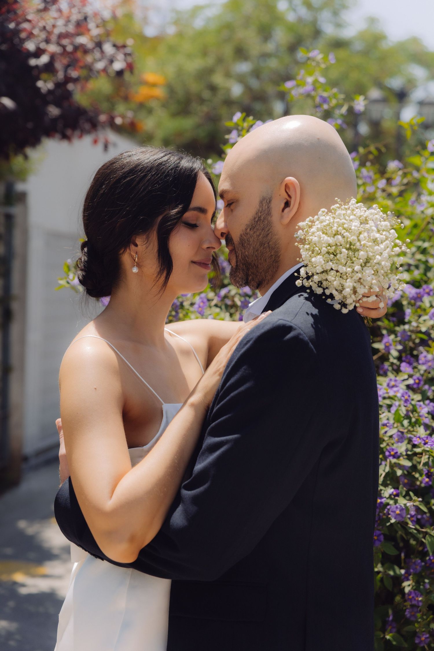 Bride in white dress and groom in dark suit nearly kissing among colorful garden flowers.