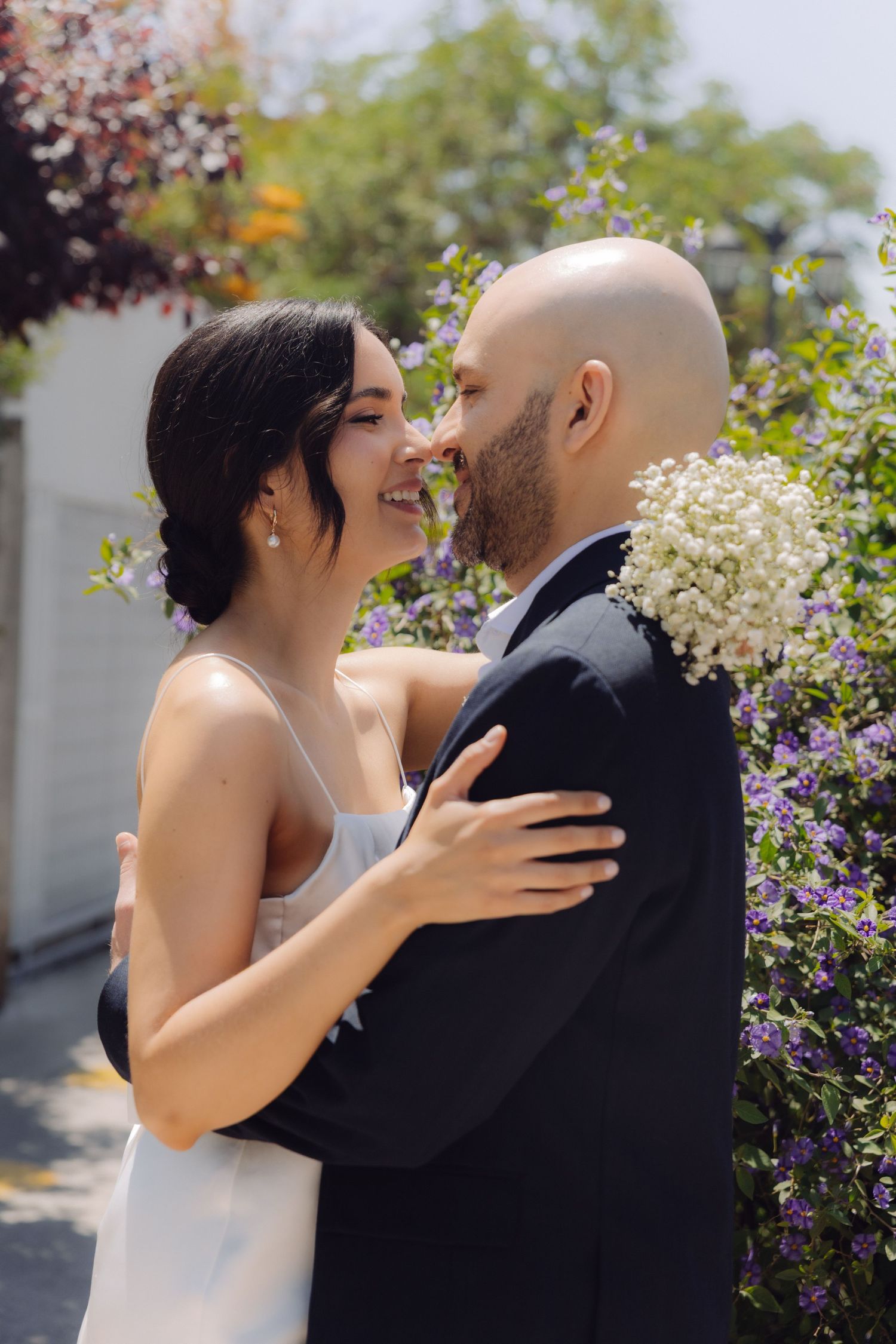 Smiling bride in white dress embracing groom in dark suit among vibrant purple garden flowers.