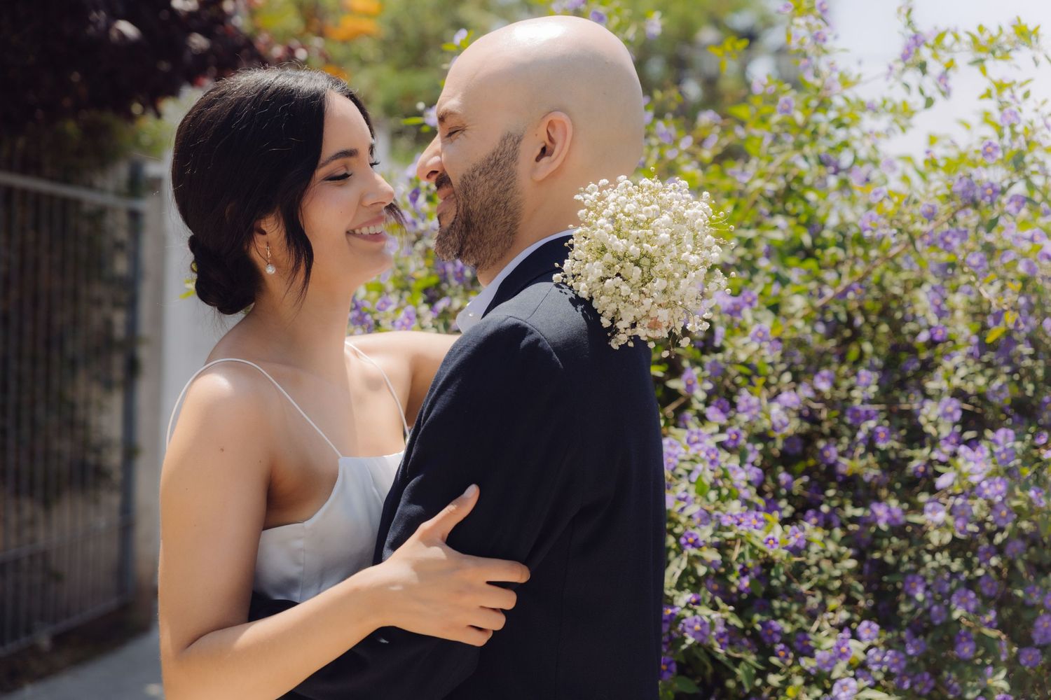 Happy bride smiling at groom while embracing outdoors with purple flowering bushes in background.