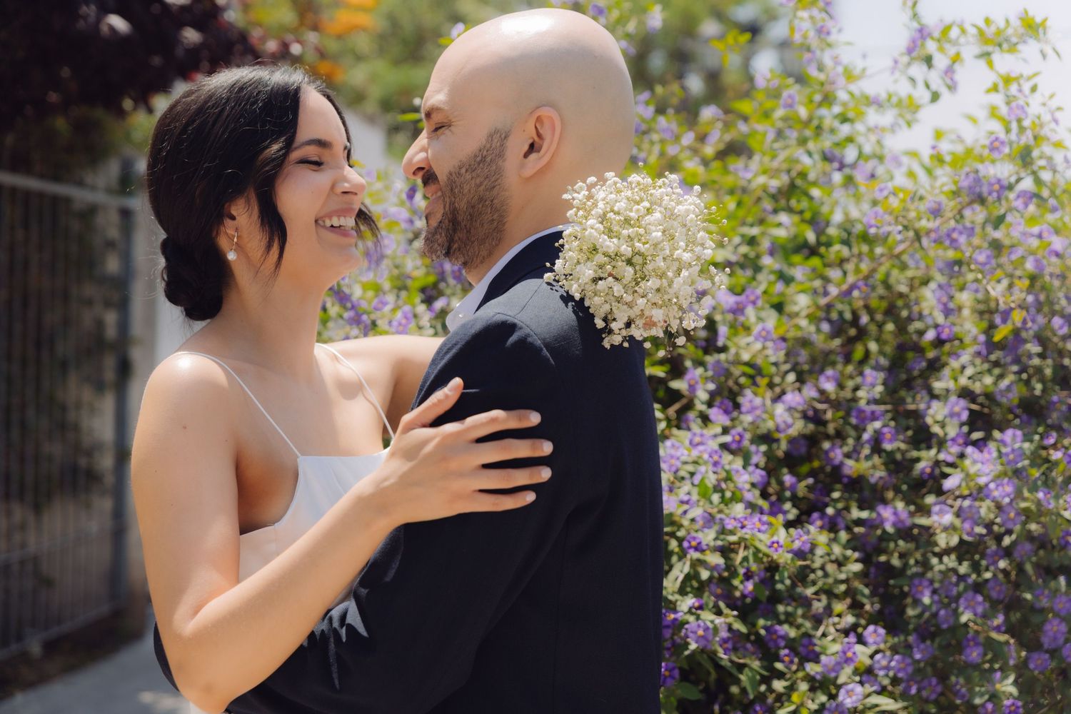 Happy bride and groom embracing outdoors near purple flowers, bride in white dress smiling at bald groom.