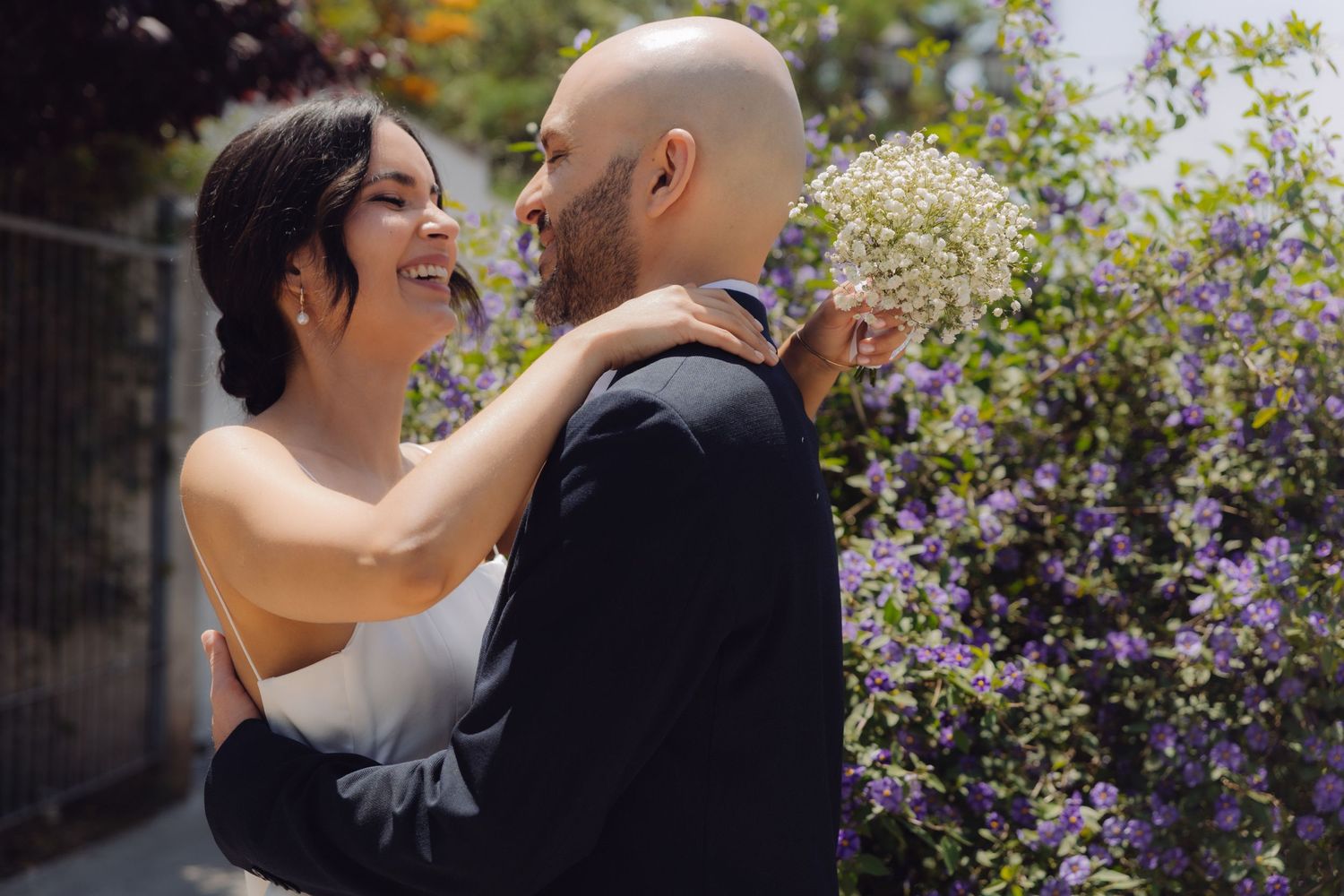 Joyful bride laughing while embracing groom holding white baby's breath in a sunny garden.