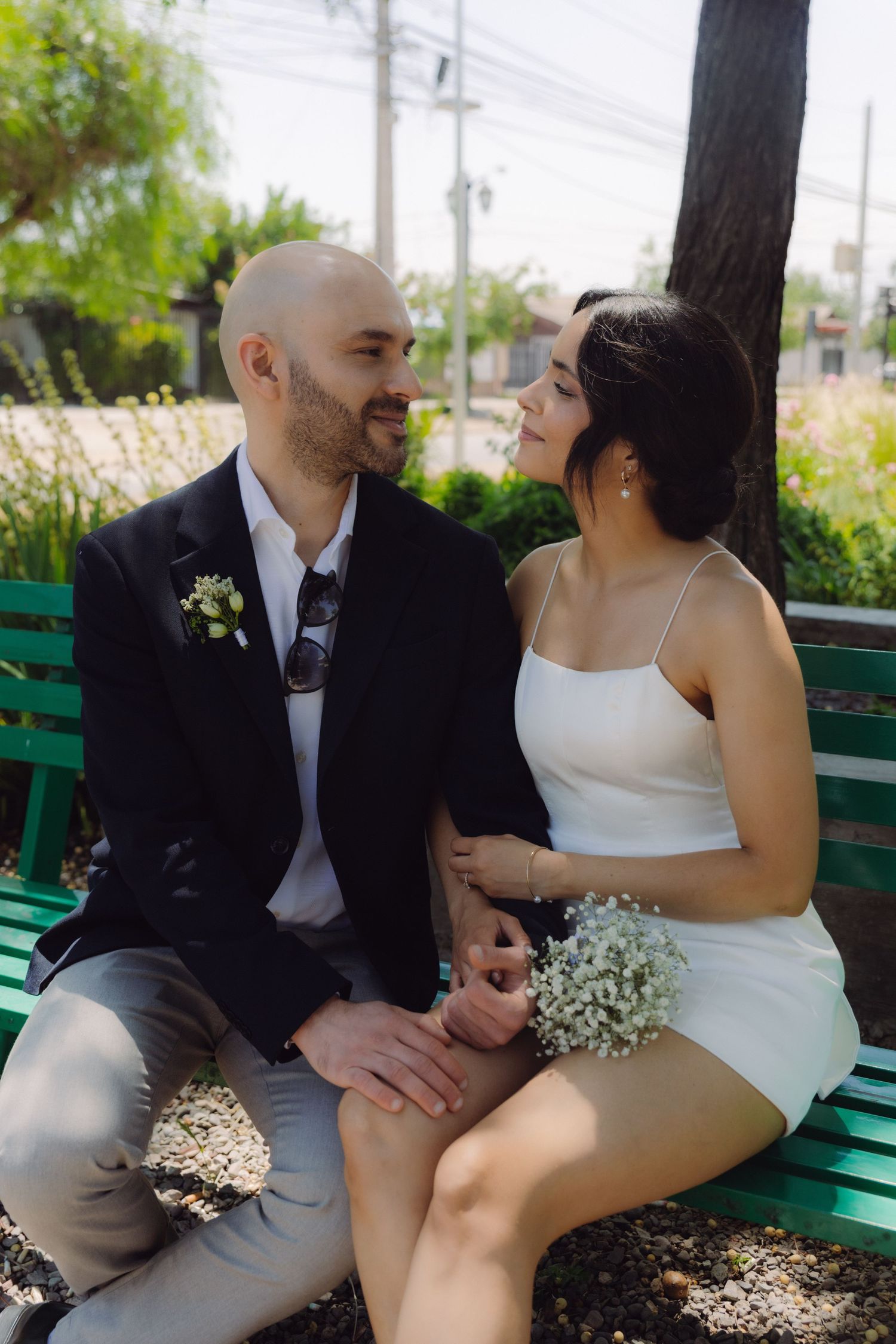 Newlywed couple sitting on a green bench holding hands and smiling at each other outdoors.