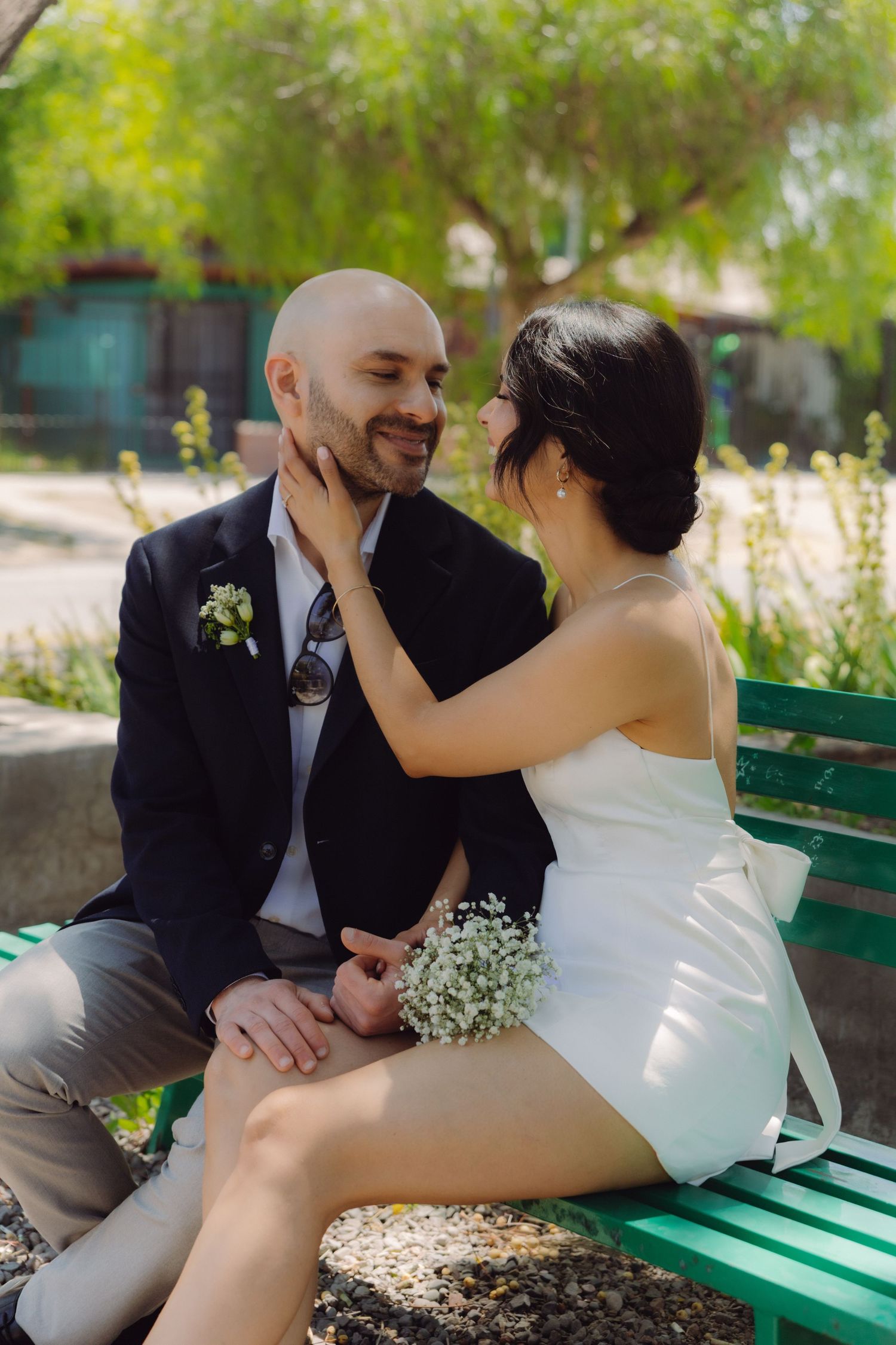 Smiling bride and groom sitting on a green bench outdoors, bride in white mini dress touching groom's face.