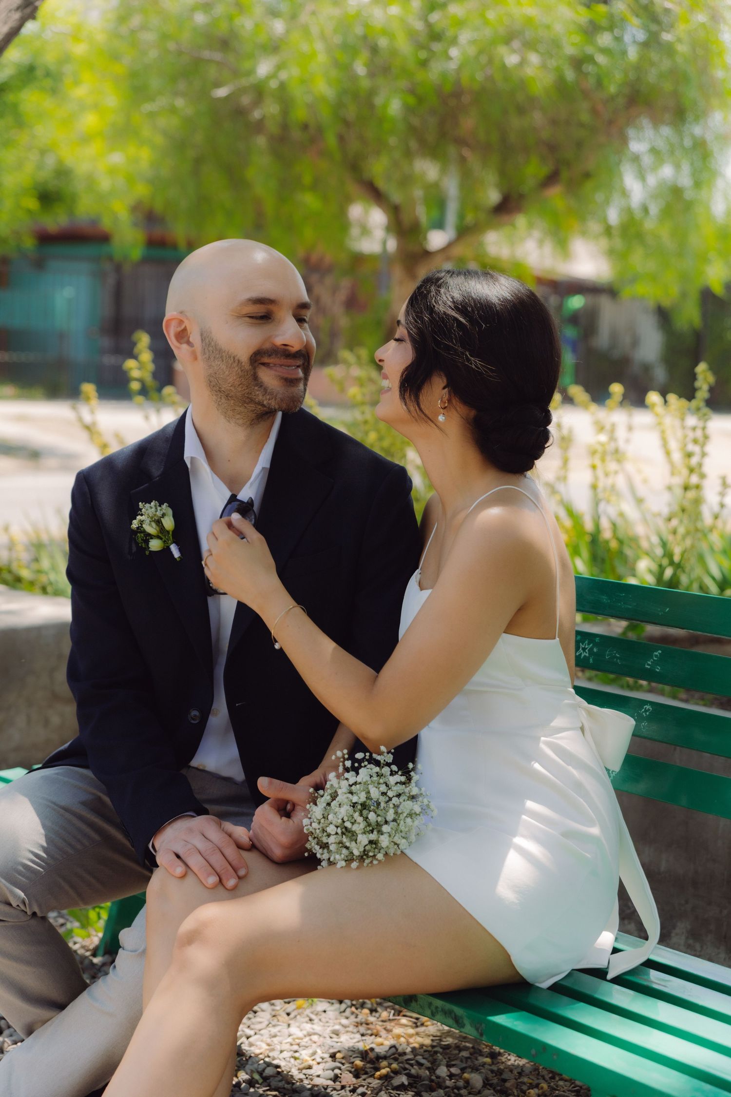 Bride in white dress adjusting groom's tie while sitting on green bench, both smiling at each other.
