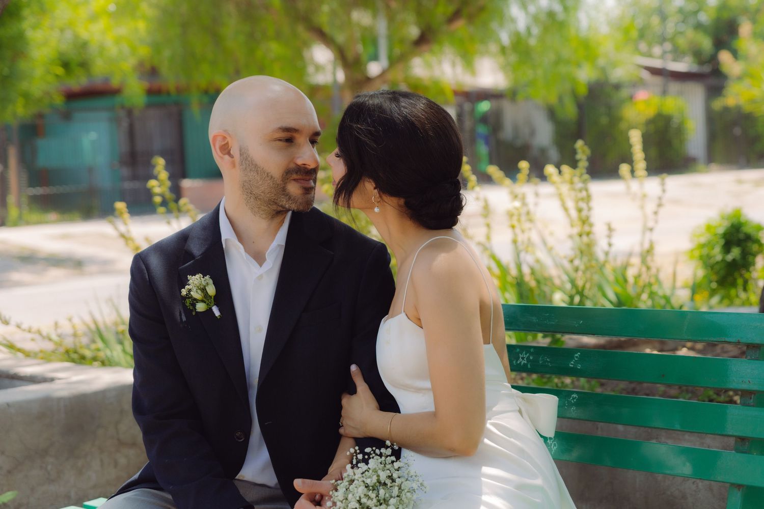 Bride and groom sitting closely on a green park bench, smiling at each other on their wedding day.