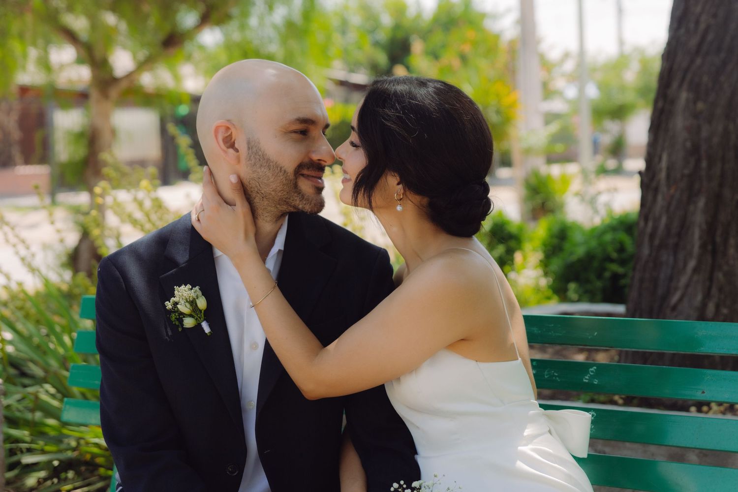 Bride tenderly holding bald groom's face on a green bench, couple sharing an intimate wedding moment outdoors.