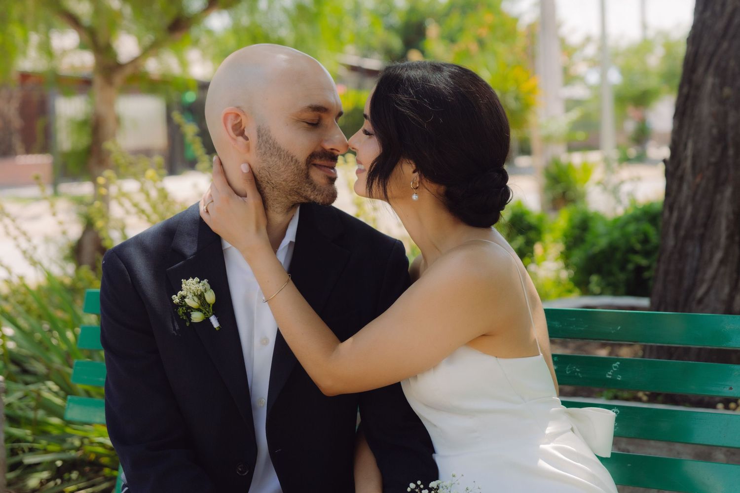Bride and bald groom in black suit sharing a romantic moment on a green bench surrounded by trees.